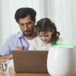 A father and his daughter sit at a table with a laptop, while HomePure Zayn ensures safe and clean indoor air